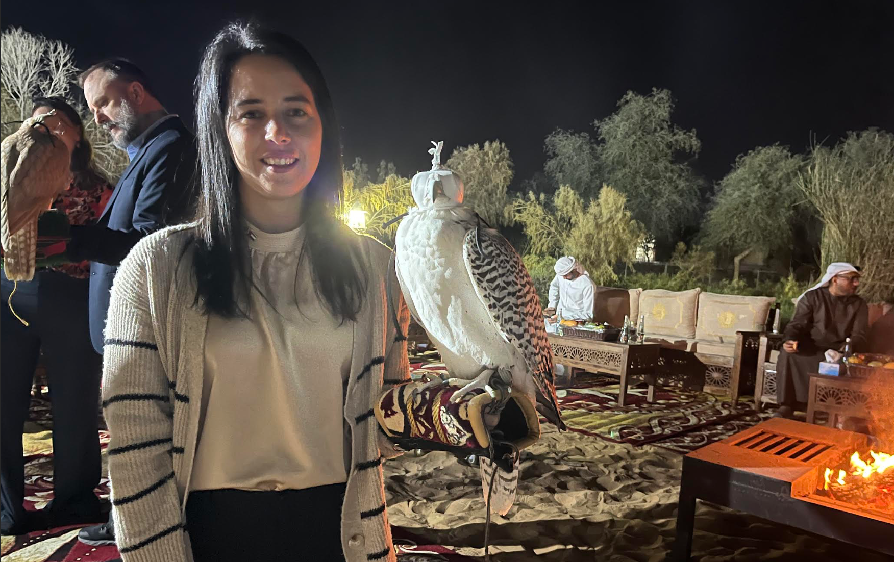Woman poses with bird perched on hand. Others in background are also holding birds and causally talking. 
